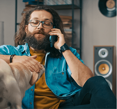 Bearded man in glasses on a couch on a phone call, petting his dog.
